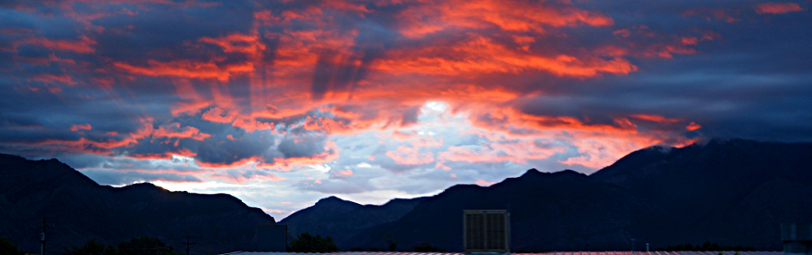 [Just above the mountain line is a series of pink-orange tinted undersides to dark blue clouds. The sun has risen over the horizon but not yet risen above the mountains. This image is several photos stitched together.]